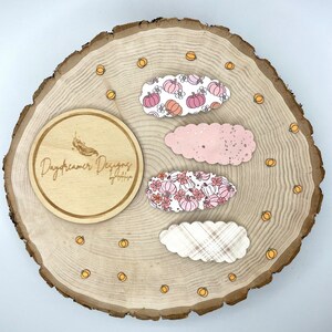 May include: Four decorative hair clips in oval shapes with fall-themed patterns. Two clips feature pumpkin and floral designs in pink and orange hues, one is solid pink, and the last has a plaid pattern. The clips are arranged on a wooden slice.
