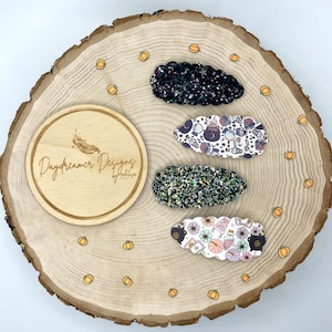 May include: Four witch-themed hair clips on a wooden background. The clips are black glitter, black and white with a witch hat and cauldron design, green glitter, and white with a potion bottle and cauldron design.