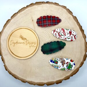 May include: Four decorative hair clips with scalloped edges are displayed on a wooden surface. The clips feature red plaid, green plaid, and Christmas-themed patterns with elves, reindeer, and presents. A wooden circle with the text "Daydreamer Designs by Alyssa" is also present.