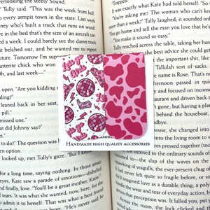 May include: Two pink and white magnetic bookmarks. One bookmark features a pink and white cow print pattern. The other bookmark features a white background with pink, white, and black illustrations of disco balls, lightning bolts, and the text "Let's Go Girls". The text "Handmade High Quality Accessories" is printed on the bottom of the bookmarks.