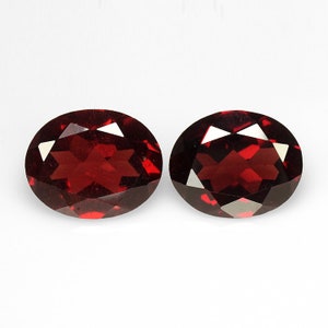 May include: Two oval-shaped, red gemstones with a faceted cut. The gemstones are set against a white background.