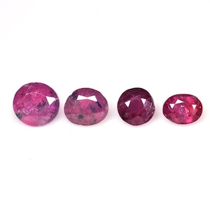 May include: Four faceted, oval and round-cut rubies in varying sizes. The gemstones display a deep red hue with visible inclusions, set against a white background. The rubies are arranged in a horizontal line, showcasing their polished surfaces.