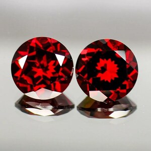 May include: Two round, red gemstones with a faceted cut. The gemstones have a dark red color and a star-like pattern in the center.