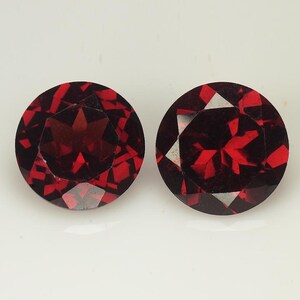 May include: Two round, faceted gemstones with a deep red color. The gems have a brilliant cut, with multiple facets reflecting light. The stones are set against a white background, highlighting their color and clarity. These gems could be used in jewelry.