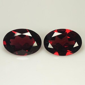 May include: Two oval-shaped, faceted gemstones with a deep red color. The gemstones are set against a white background.