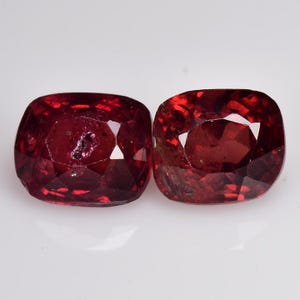 May include: Two cushion-cut gemstones with a deep red hue. The gems are highly faceted, reflecting light and exhibiting a high degree of clarity. The stones are displayed against a white backdrop.