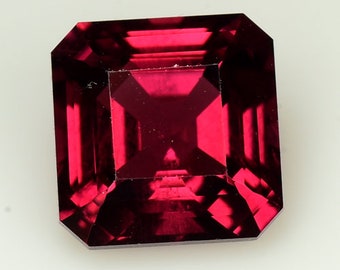 May include: A deep red gemstone with a square emerald cut. The gemstone is faceted and has a high polish.