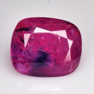 Puede incluir: Una gema de rubí tallada en cojín. La gema muestra un tono rojo rosado profundo y saturado con inclusiones internas y una superficie pulida. El corte presenta múltiples facetas que reflejan la luz, creando brillo.