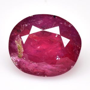 Può includere: Una gemma sfaccettata di forma ovale con una tonalità rosso intenso. La gemma presenta sfaccettature triangolari bianche nella parte superiore e una superficie leggermente irregolare. La pietra è posta su uno sfondo bianco, evidenziando il suo colore e la sua chiarezza.