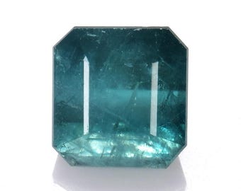 May include: A teal-colored gemstone with an emerald cut. The gem has a rectangular shape with beveled edges and two vertical, parallel facets. The stone is translucent, with internal inclusions visible. The gem is set against a white background.