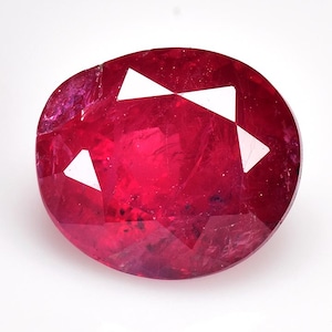Può includere: Pietra preziosa di rubino taglio ovale con una tonalità rosso intenso. La gemma presenta sfaccettature che riflettono la luce, creando forme triangolari bianche brillanti. Il rubino è posto su uno sfondo bianco semplice, evidenziando il suo colore e la sua chiarezza.