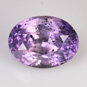 Peut inclure: Une pierre pr&eacute;cieuse ovale et facett&eacute;e, de couleur violet clair. La gemme pr&eacute;sente des inclusions sombres dispers&eacute;es, ajoutant une texture unique. Les facettes cr&eacute;ent un jeu de lumi&egrave;re, rehaussant l'&eacute;clat de la pierre. Vue rapproch&eacute;e.