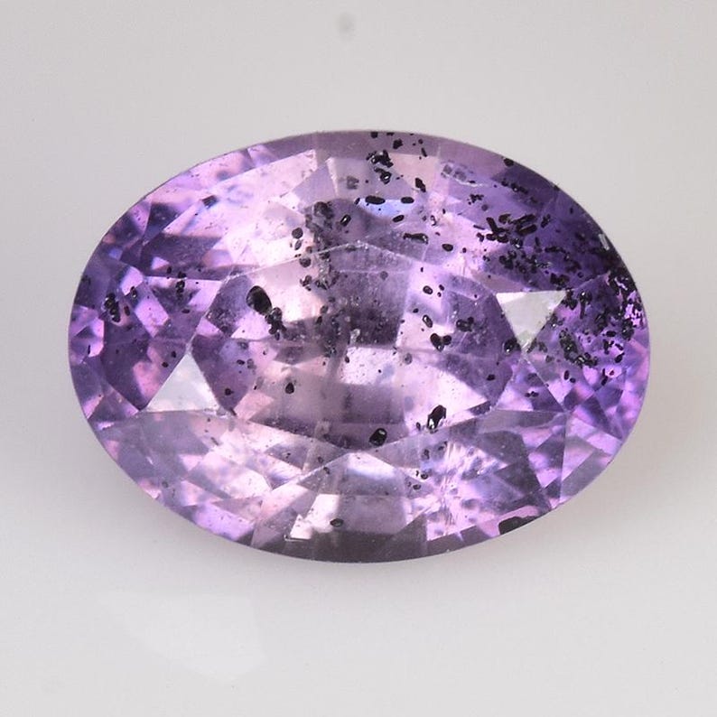 Peut inclure: Une pierre pr&eacute;cieuse ovale et facett&eacute;e, de couleur violet clair. La gemme pr&eacute;sente des inclusions sombres dispers&eacute;es, ajoutant une texture unique. Les facettes cr&eacute;ent un jeu de lumi&egrave;re, rehaussant l'&eacute;clat de la pierre. Vue rapproch&eacute;e.