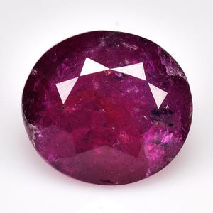 Op de afbeelding: Een gefacetteerde, ovaalvormige robijn edelsteen. De edelsteen heeft een diepe, rijke magenta kleur met witte driehoekige facetten. Het oppervlak van de robijn heeft een licht ruwe textuur. De edelsteen staat tegen een witte achtergrond.