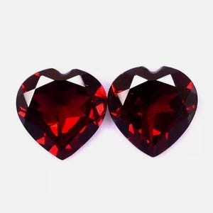 May include: Two heart-shaped, faceted, deep red gemstones. The gems have a brilliant cut with multiple facets that reflect light. The heart shape is clearly defined, with a small indentation at the top. The gems are set against a white background.
