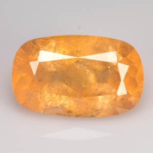 Pode incluir: Uma gema facetada em forma oval com um tom quente de laranja dourado. A gema apresenta uma série de facetas angulares que refletem a luz, criando um efeito brilhante. A pedra está sobre um fundo liso e claro.