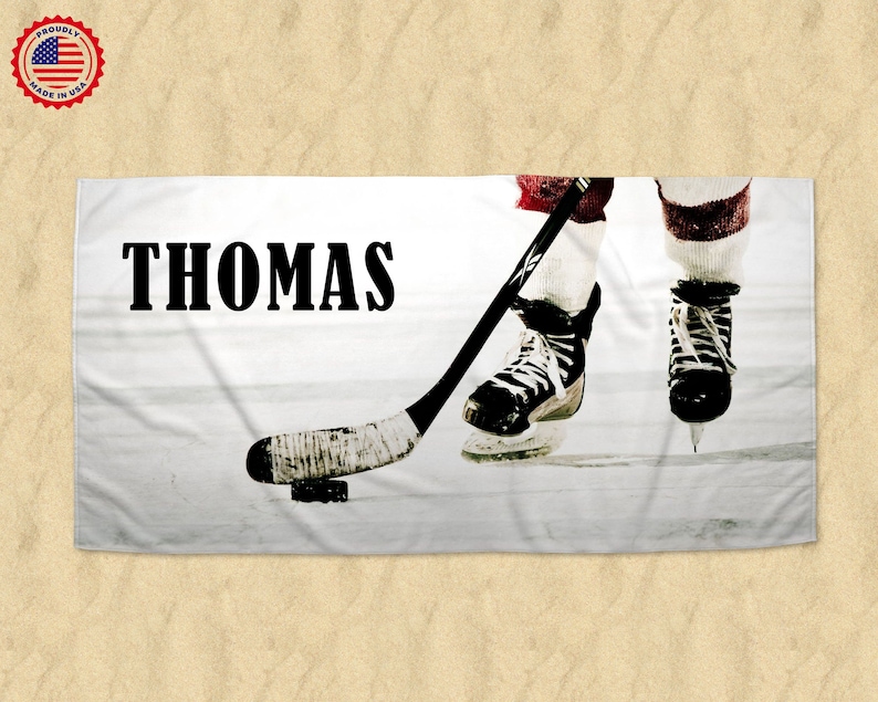 K&ouml;nnte beinhalten: Ein wei&szlig;es Strandtuch mit den Beinen eines Hockeyspielers und einem Hockeyschl&auml;ger. Der Spieler tr&auml;gt schwarz-wei&szlig;e Schlittschuhe und rot-wei&szlig;e Socken. Der Name "THOMAS" ist in schwarzen Buchstaben auf dem Handtuch gedruckt.
