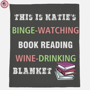 May include: A gray throw blanket with the text "This is Katie's Binge-Watching Book Reading Wine-Drinking Blanket" in white, pink, and green. A stack of three books is in the bottom right corner.