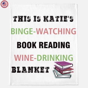 May include: White blanket with the text "This is Katie's Binge-Watching Book Reading Wine-Drinking Blanket" in black. There are three stacked books with purple, red, and white covers in the bottom right corner.