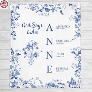 Op de afbeelding: Witte deken met een blauw bloemenpatroon en de tekst "God Says I Am ANNE". De letters A, N, N en E zijn gekoppeld aan de woorden Adorned, Nurtured, Near en Eternal, met bijbehorende bijbelverzen.