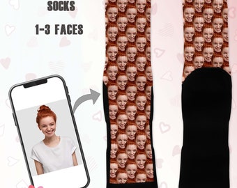 May include: A pair of custom face mashup socks. The socks feature a repeating pattern of a person's face, with a black heel and toe. The image also shows a smartphone displaying the face used for the socks. Text on the image reads "FACE MASHUP SOCKS" and "1-3 FACES".