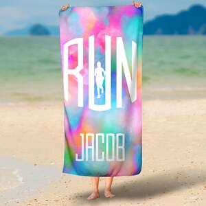 以下が含まれることがあります： 「RUN」の白いデザインが施されたカラフルなビーチタオル。ランニングする人物と、その下に「JACOB」という名前が書かれています。