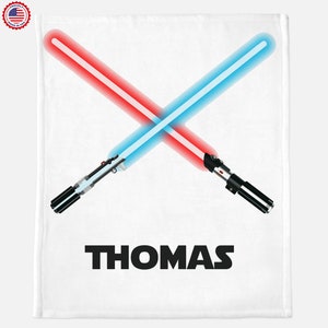Puede incluir: Manta blanca con sables de luz rojos y azules cruzados. El nombre "THOMAS" está impreso en letras mayúsculas negras en negrita debajo. Un logotipo de "Proudly Made in USA" está en la esquina superior izquierda.