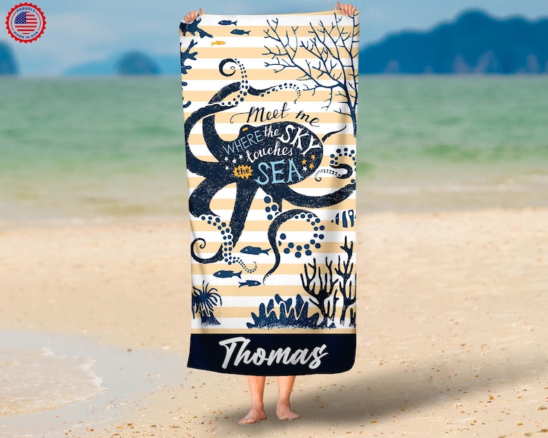 Pode incluir: Uma toalha de praia com um desenho de polvo azul marinho e o texto "Meet me where the sky touches the sea". A toalha tem um fundo listrado com ilustra&ccedil;&otilde;es de corais e peixes. O nome "Thomas" &eacute; impresso na parte inferior.