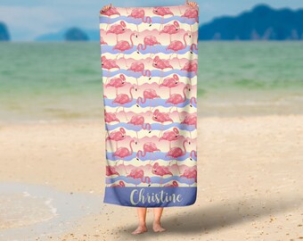 Op de afbeelding: Een strandlaken met een herhaald patroon van roze flamingo's op een crèmekleurige achtergrond met blauwe en roze golfstrepen. De onderste rand is blauw met de naam "Christine" in witte letters.