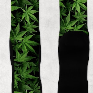 Op de afbeelding: Sokken met een herhaald patroon van groene cannabisbladeren op een zwarte achtergrond. De sokken hebben zwarte hielen en tenen. Het ontwerp bedekt de hele voorkant van de sokken.