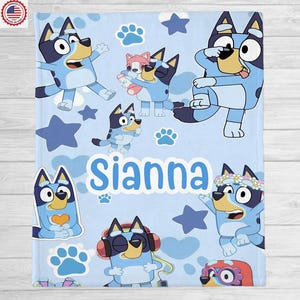 May include: A light blue blanket featuring cartoon characters, paw prints, and stars. The name "Sianna" is displayed in large, light blue letters. The blanket has a soft, plush appearance, suitable for children.