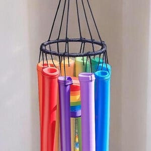 May include: A hanging wind chime featuring multiple cylindrical tubes in vibrant colors including red, orange, yellow, green, blue, and purple. The tubes are suspended from a black circular frame by black cords, topped with a rainbow-striped bead.