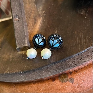 Könnte beinhalten: Ein Paar handgefertigte Ohrstecker. Jeder Ohrring hat eine schwarze, kreisförmige Basis mit einem hellblauen Blumendesign und ist mit kleinen, dunkelblauen und klaren Perlen verziert. Eine große, irisierende weiße Perle hängt von jedem Ohrring.