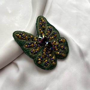 Puede incluir: Broche de mariposa verde hecho a mano con intrincados abalorios en tonos amarillos, morados y verdes. El centro presenta un gran cristal iridiscente. El broche probablemente está hecho de tela y abalorios, y es un accesorio decorativo.