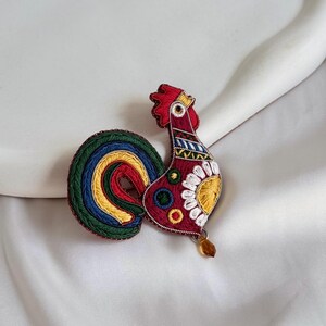 May include: Embroidered rooster brooch with a red body, yellow and white accents, and a colourful tail. The brooch has a red comb and a small, dangling amber-coloured bead. The brooch is set against a white background.