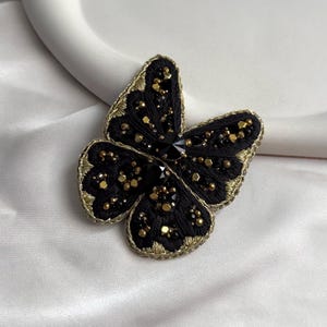 Puede incluir: Broche de mariposa negra con ribete y detalles dorados. Las alas están bordadas con hilo negro y adornadas con cuentas doradas y negras. Una gran gema negra se encuentra en el centro de la mariposa.
