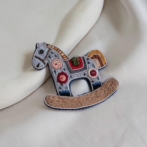 May include: A detailed brooch depicting a rocking horse. The grey horse features embroidered patterns in red, brown, and green. The rocking horse has a brown base and a colourful saddle. A unique accessory, perfect for adding a touch of whimsy.
