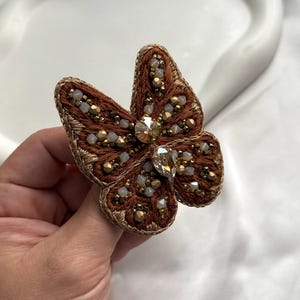 Puede incluir: Broche de mariposa marrón con intrincados bordados y abalorios. Las alas son de color marrón intenso, delineadas con hilo dorado. El cuerpo presenta cristales transparentes y brillantes y pequeñas cuentas doradas e iridiscentes.