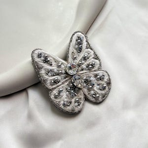 Puede incluir: Broche de mariposa hecho a mano con detalles blancos, plateados y cristales transparentes. Las alas están bordadas con hilo plateado y adornadas con cuentas y cristales. El centro presenta un gran cristal brillante.