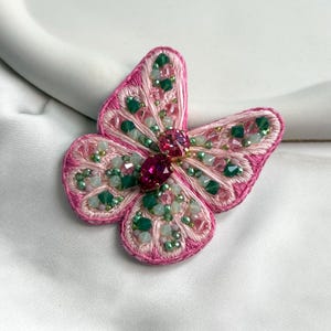 Puede incluir: Broche de mariposa bordada en rosa con detalles intrincados. Las alas están delineadas con hilo rosa y adornadas con cuentas verdes y rosas. Una gran gema rosa facetada está en el centro. Un accesorio hecho a mano para un toque caprichoso.