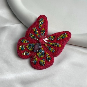 Puede incluir: Broche de mariposa roja con alas bordadas, adornada con cuentas de colores y dos grandes gemas facetadas. Un accesorio vibrante para añadir un toque de elegancia.