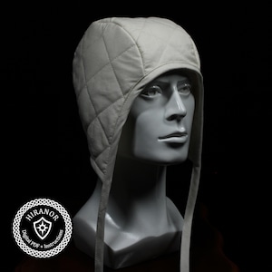 May include: A light gray quilted hat with ear flaps and ties, displayed on a mannequin head. The hat features a diamond pattern and is made of a soft fabric. The background is black.