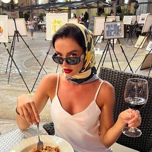 Może przedstawiać: Kobieta w białym topie na ramiączkach i ż&oacute;łto-czarnym wzorzystym szaliku siedzi przy stole z talerzem spaghetti i szklanką czerwonego wina. Ma na sobie okulary przeciwsłoneczne i czerwony manicure.
