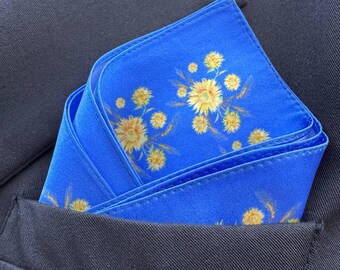 May include: A blue pocket square with a floral pattern of yellow flowers and wheat stalks. The pocket square is folded and placed in a suit jacket pocket.