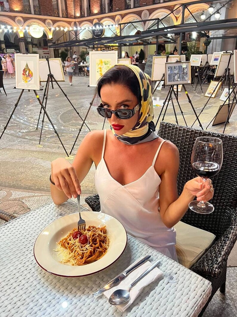Może przedstawiać: Kobieta w białym topie na ramiączkach i ż&oacute;łto-czarnym wzorzystym szaliku siedzi przy stole z talerzem spaghetti i szklanką czerwonego wina. Ma na sobie okulary przeciwsłoneczne i czerwony manicure.