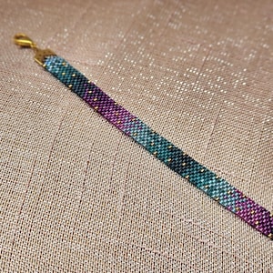 Peut inclure: Un bracelet de perles avec un dégradé de bleu sarcelle, de violet et d'or. Le bracelet est composé de petites perles espacées uniformément et possède un fermoir doré. Le bracelet est présenté sur une surface texturée de couleur beige.