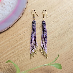 Peut inclure: Une paire de longues boucles d'oreilles en perles avec un dégradé de perles violettes, roses et dorées. Les boucles d'oreilles sont faites de petites perles carrées et ont des crochets dorés.