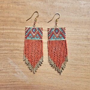 Könnte beinhalten: Ein Paar orangefarbene und türkisfarbene Perlenohrringe mit goldenen Akzenten. Die Ohrringe haben ein geometrisches Muster und ein Fransen-Design.