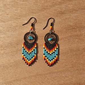 May include: A pair of beaded earrings featuring a turquoise stone and a black dreamcatcher design. The earrings are made with black, blue, red, white, and yellow beads.