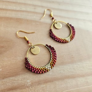 Peut inclure: Une paire de boucles d'oreilles créoles dorées avec un motif de perles. Les perles sont dans des tons de rouge, orange et blanc. Chaque boucle d'oreille a un petit charme rond en or.